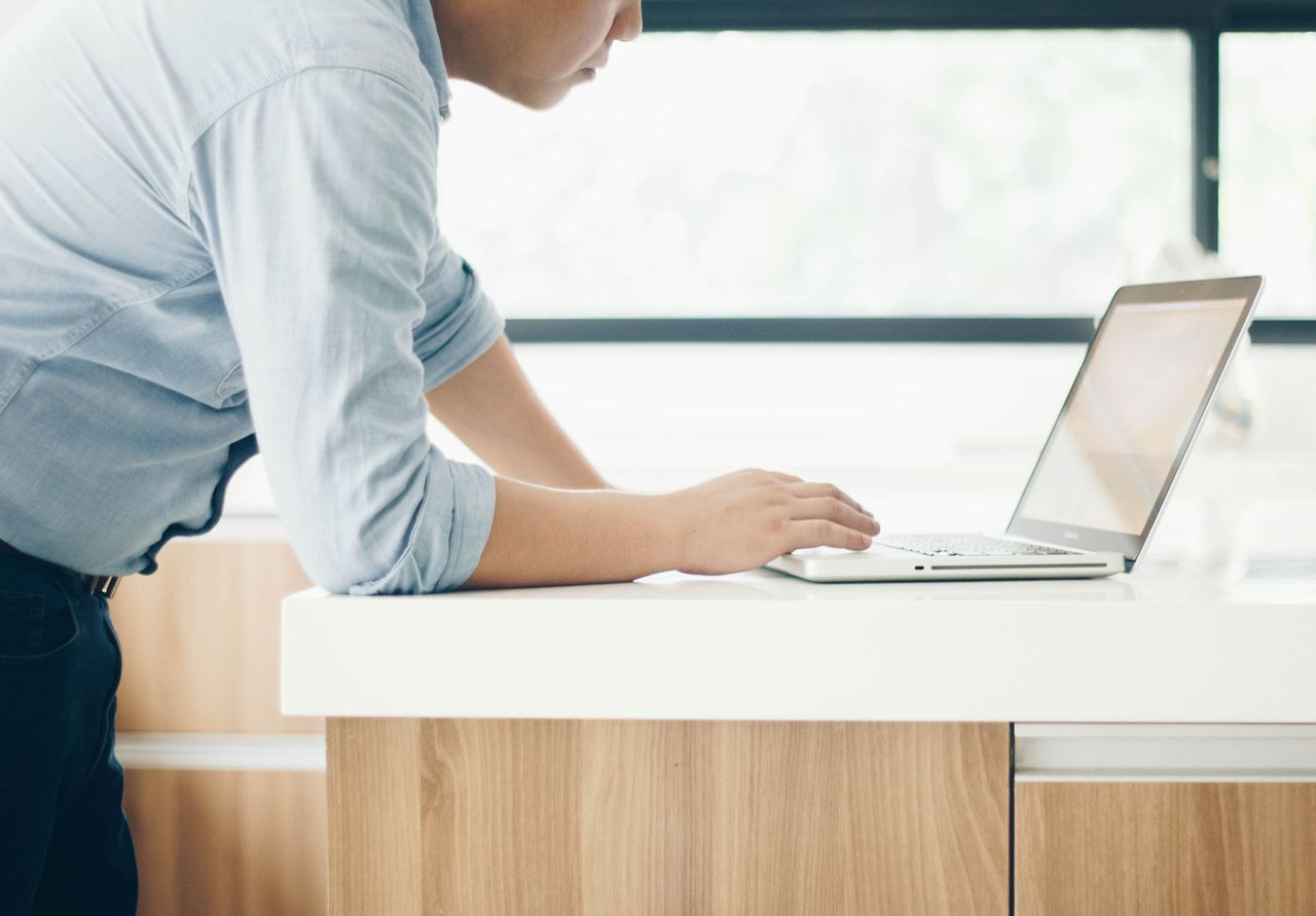 A man leaning on a desk working on a laptop in a bright indoor office setting.